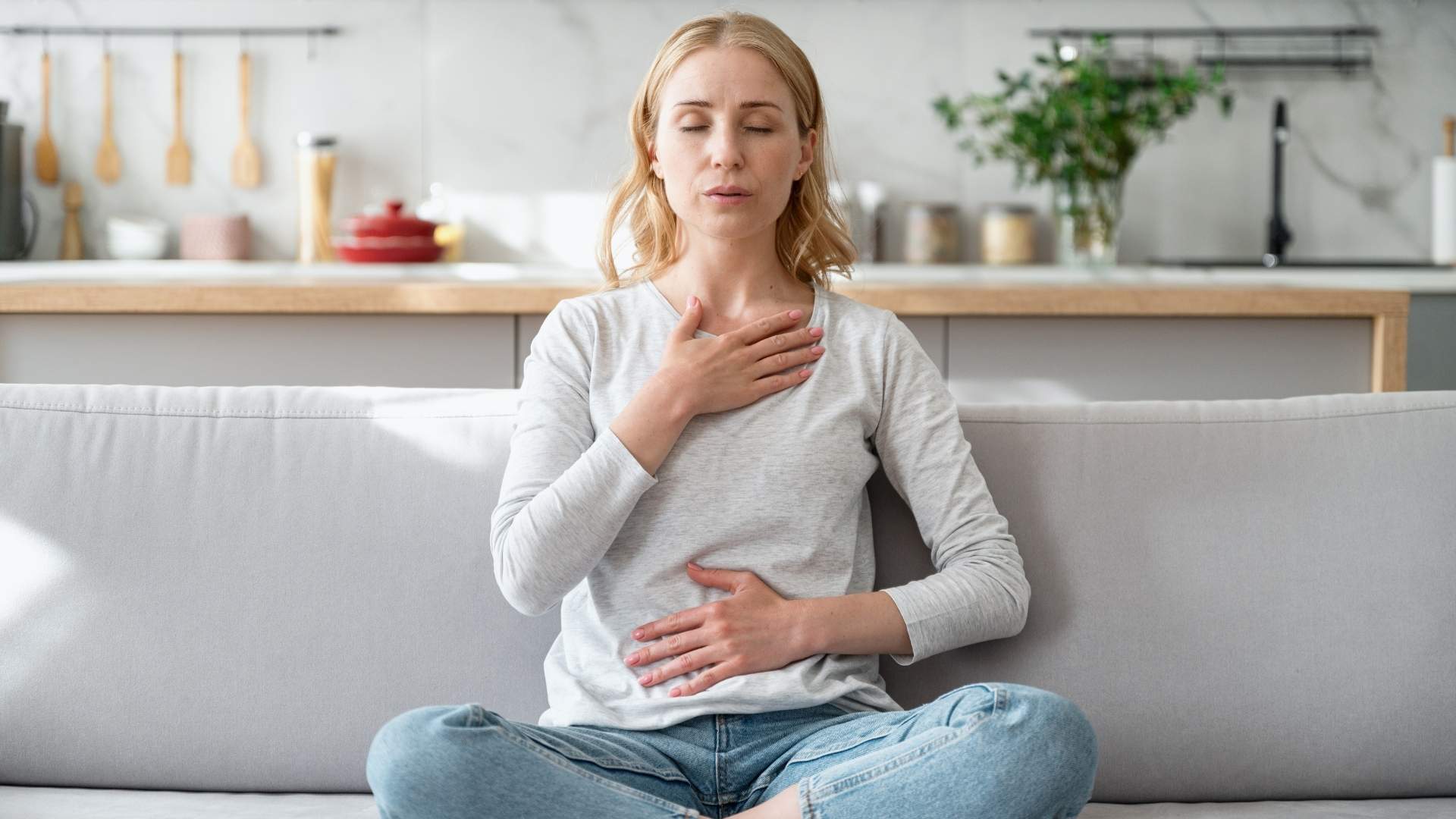 Mujer sentada en un sofá, con las manos sobre el estómago, reflexionando sobre rituales de transformación.